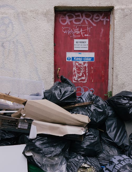 Exterior scene featuring a red metal door with graffiti, a white 'Keep Clear' sign, and a 'No Money' notice. In front of the door, there are numerous black and some cardboard garbage bags, along with 