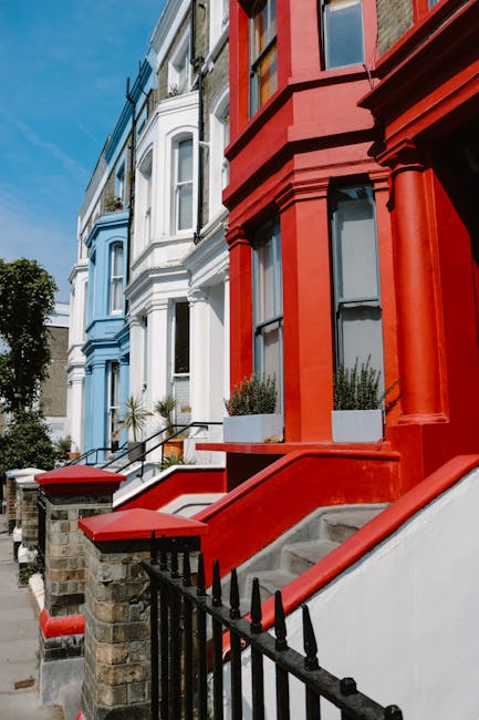 Exterior view of a row of Victorian-style residential buildings with bay windows and decorative architecture in bright blue, white, and red colors. The buildings feature stoops with concrete steps, bl