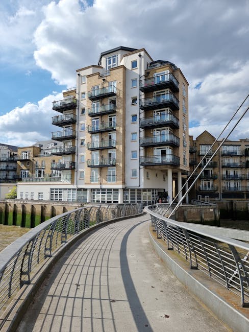 Image of a riverside apartment building in Shad Thames viewed from the river, with a paved promenade and green garden area illuminated by exterior lights, surrounded by a wooden and metal railing. The
